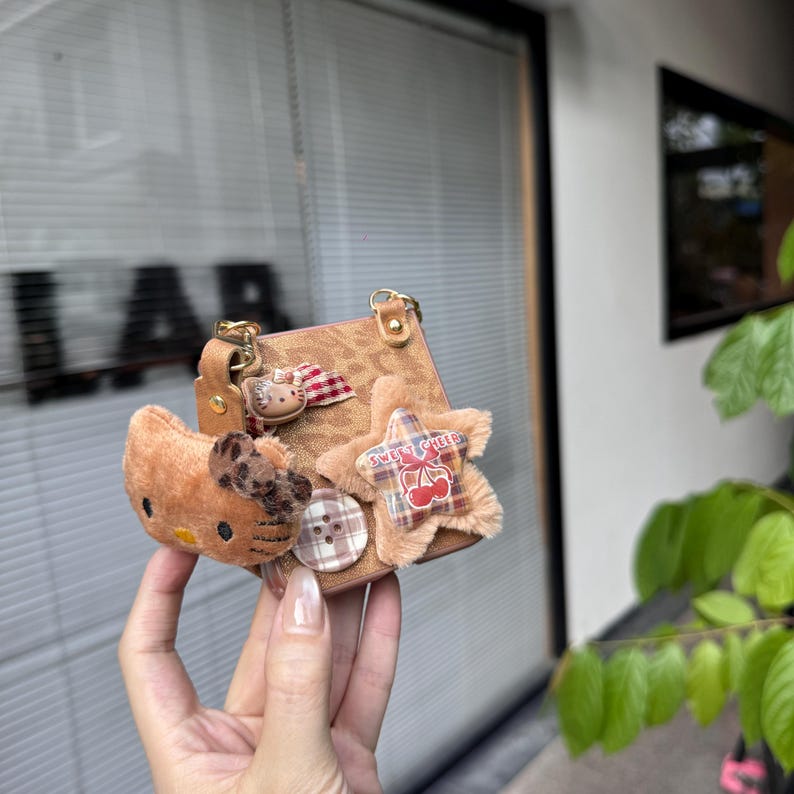 May include: A small, brown handbag with a gold chain strap, adorned with a plush Hello Kitty, a star-shaped patch with "Sweet Cherry" text, and a button. The bag has a textured surface and a small handle.
