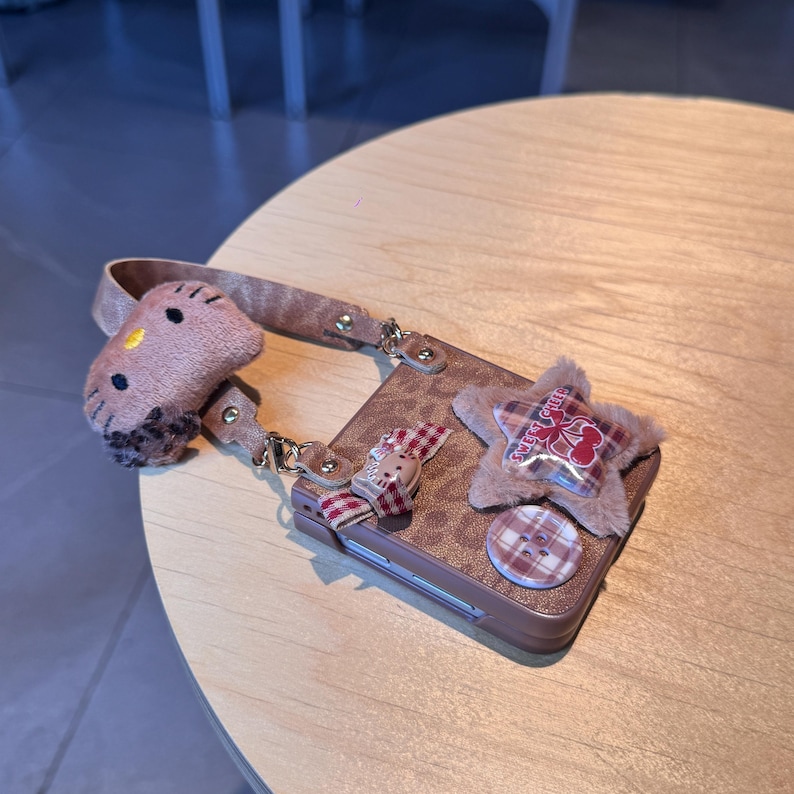 May include: A brown phone case with a leopard print design, adorned with a plush star, a Hello Kitty charm, and a plaid bow. The case has a matching strap and a button detail. The overall aesthetic is cute and girly.