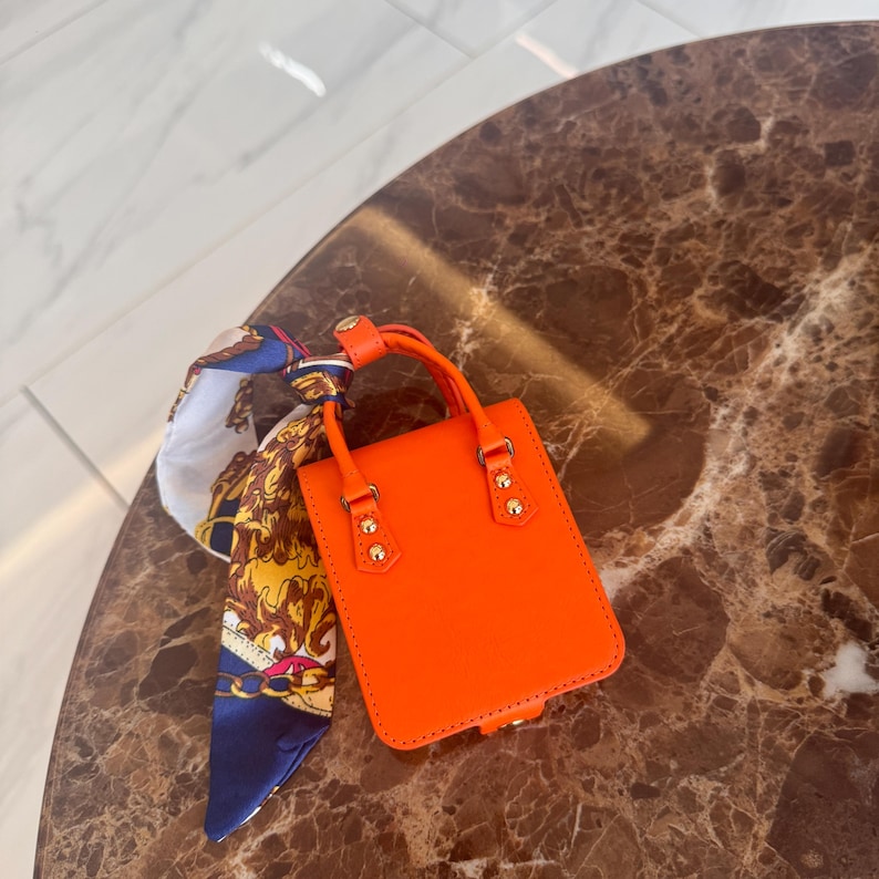 May include: Bright orange mini handbag with a top handle and gold-tone hardware. A patterned scarf in blue, gold, and white is tied to the handle. The bag has a rectangular shape and a structured design.