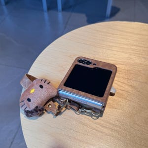 May include: A brown leather phone case with a black screen and a small, plush Hello Kitty charm. The case has a silver hinge and a strap. The phone case is on a light brown wooden surface.