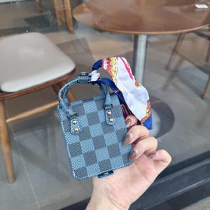 May include: A small, blue and gray checkered handbag with a matching handle and gold-colored accents. A colorful scarf is tied to the handle. The bag is held in a person's hand.