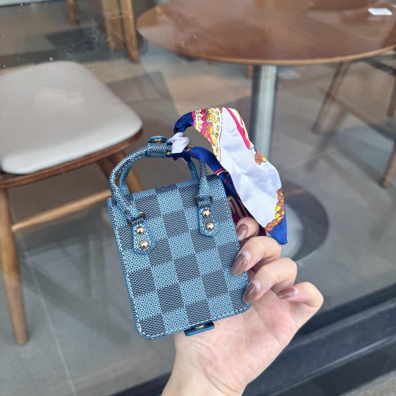May include: A small, blue and gray checkered handbag with a matching handle and gold-colored accents. A colorful scarf is tied to the handle. The bag is held in a person's hand.