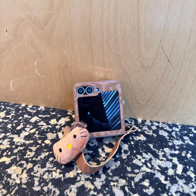 May include: A brown phone case with a matching horse-shaped charm. The phone case has a textured appearance and a camera cutout. The horse charm is light brown with a yellow nose and a brown leather strap.