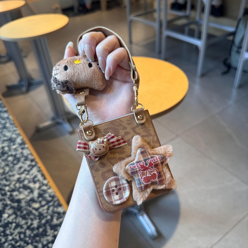 May include: Brown phone case with a detachable strap, adorned with a plush star, a Hello Kitty bow, and a button. The case features a small plush animal charm and gold-colored hardware.
