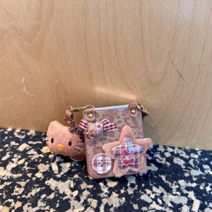 May include: A small, brown purse with a leopard print pattern, adorned with a plush star, a Hello Kitty bow, and a button. A Hello Kitty plush keychain hangs from the purse. The star has the words "Sweet Cherry" on it.