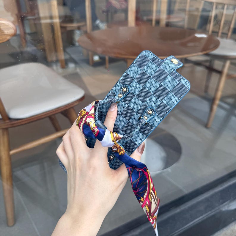 May include: Blue and black checkered phone case with a matching strap and gold-colored hardware. The case is paired with a colorful patterned scarf. The phone case is designed to protect the phone from scratches and damage.