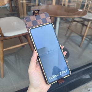 May include: Brown and gold phone case for a Galaxy Z Flip7. The case features a checkered pattern on the flap and a gold-colored frame. The phone screen displays the text "Galaxy Z Flip7".