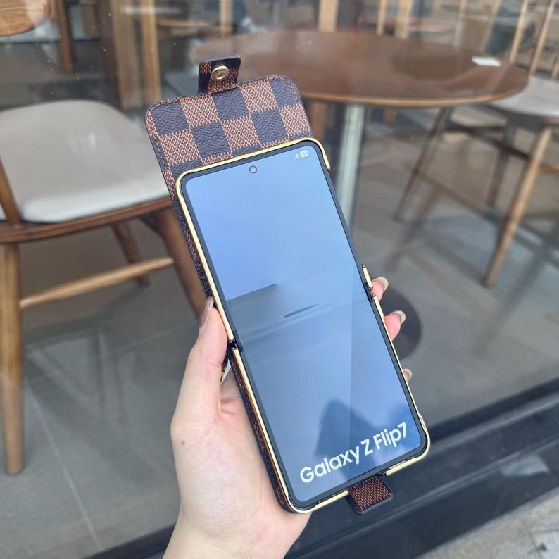 May include: Brown and gold phone case for a Galaxy Z Flip7. The case features a checkered pattern on the flap and a gold-colored frame. The phone screen displays the text "Galaxy Z Flip7".