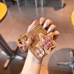 May include: Brown phone case with a leopard print design, adorned with a plush star, a button, and a Hello Kitty charm. The star-shaped embellishment has the text "SWEET CHEER" and a cherry design. The case has a gold-colored metal clasp.