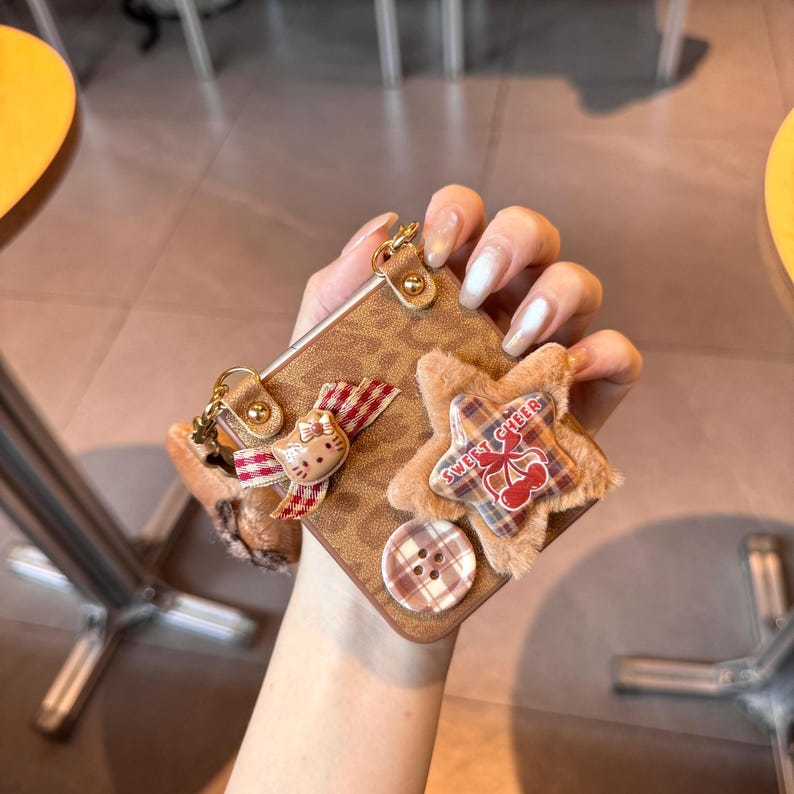 May include: Brown phone case with a leopard print design, adorned with a plush star, a button, and a Hello Kitty charm. The star-shaped embellishment has the text "SWEET CHEER" and a cherry design. The case has a gold-colored metal clasp.