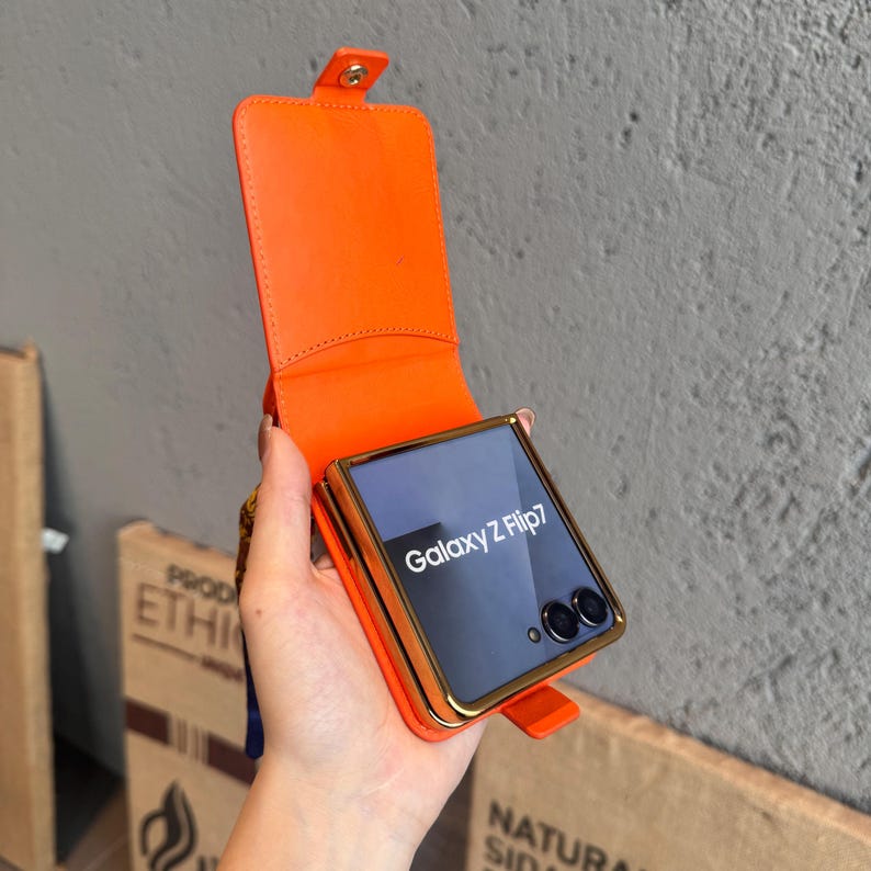 May include: Orange leather phone case for a Galaxy Z Flip7. The case has a gold-colored frame and a matching orange flap that secures with a snap. The phone screen displays the words "Galaxy Z Flip7".