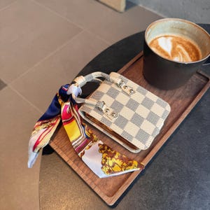May include: A small, cream-colored handbag with a checkered pattern and gold-colored hardware. A colorful scarf is tied to the handle. The bag sits on a wooden tray with a cup of coffee.
