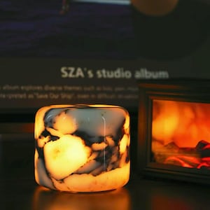 May include: A cylindrical lamp with a smooth, translucent white surface featuring dark, irregular marble-like veining. The lamp emits a warm, glowing orange light from within, illuminating the stone-like texture and creating a soft, ambient effect.