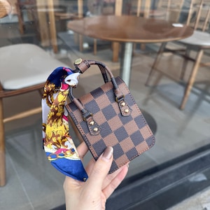 May include: A small, brown and tan checkered handbag with a matching handle and gold-colored hardware. A colorful scarf with a blue, white, and gold pattern is tied to the handle. The bag is held in a person's hand.