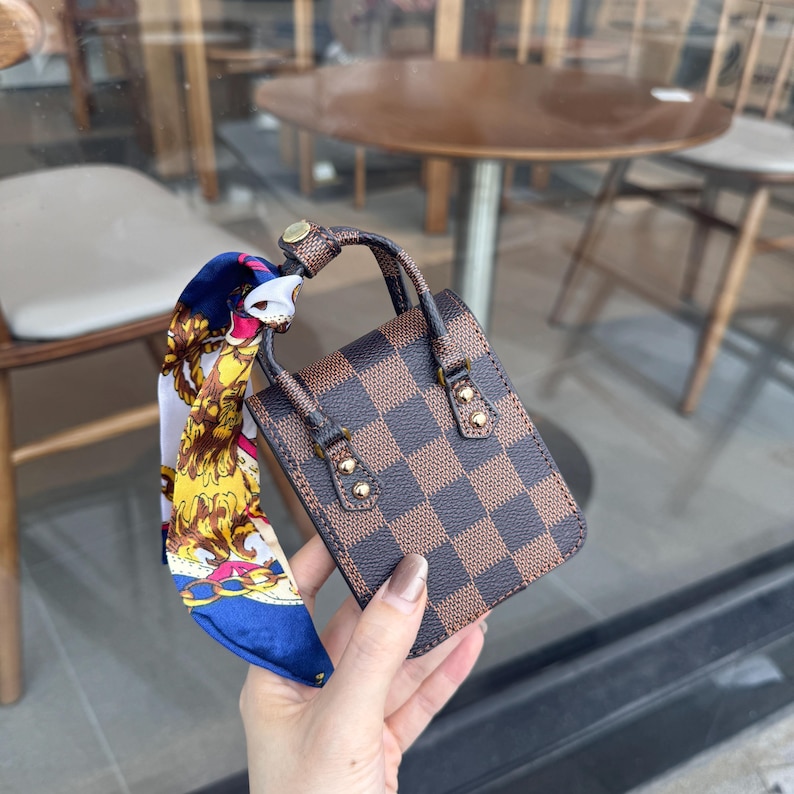 May include: A small, brown and tan checkered handbag with a matching handle and gold-colored hardware. A colorful scarf with a blue, white, and gold pattern is tied to the handle. The bag is held in a person's hand.