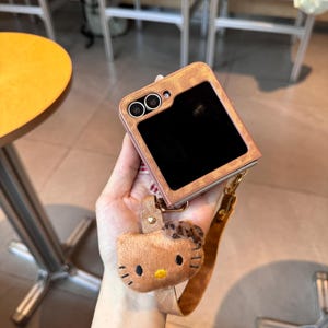 May include: A brown and pink phone case with a black screen and two camera lenses. Attached is a brown Hello Kitty plush keychain with a leopard print bow and a matching wrist strap. The case has a textured, metallic finish.