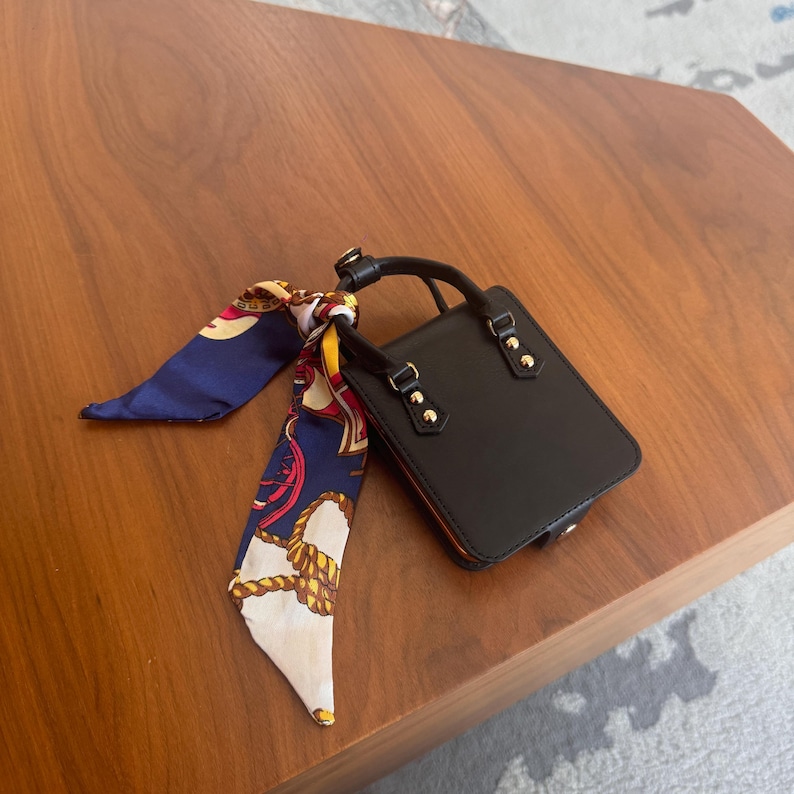 May include: A small, black handbag with a top handle and gold-colored hardware. A patterned silk scarf in navy blue, white, and gold is tied to the handle. The bag is rectangular and appears to be made of leather.