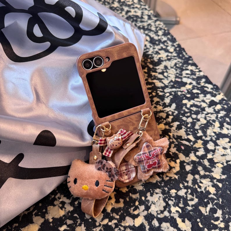 May include: Brown phone case with a flip design, featuring a plush Hello Kitty charm and a star-shaped accessory. The accessories include a red and white plaid bow and the words "Sweet Cheer".