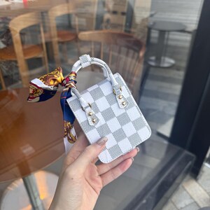 May include: A small white and gray checkered handbag with a short handle and gold-colored hardware. A colorful scarf is tied to the handle. The bag is held in front of a blurred background.