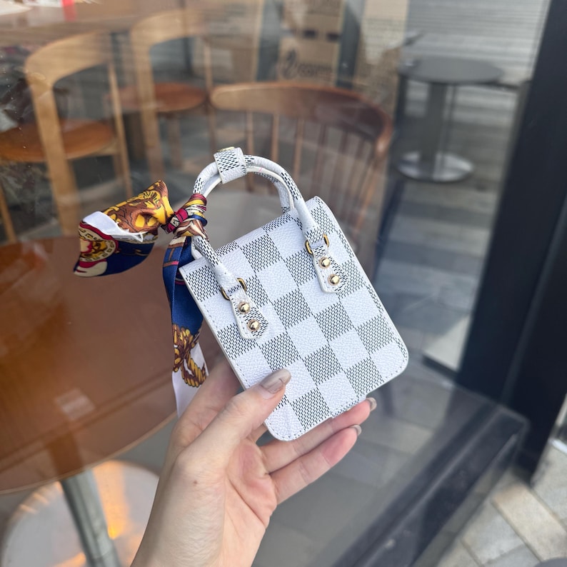 May include: A small white and gray checkered handbag with a short handle and gold-colored hardware. A colorful scarf is tied to the handle. The bag is held in front of a blurred background.