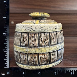 May include: A barrel-shaped container with a lid, featuring a brown wood-grain texture and yellow bands. A clear measuring cup filled with blue liquid is positioned next to it, with a black ruler showing a height of approximately 3.5 inches.