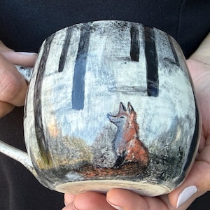 May include: A ceramic mug featuring a hand-painted scene of a fox sitting in a forest with tall, dark tree trunks against a light, textured background. The fox is depicted in shades of orange and brown, with a long, bushy tail.