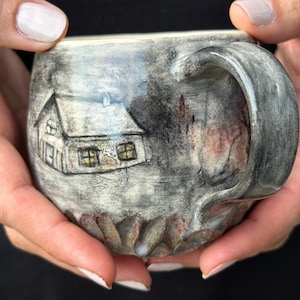 May include: A ceramic mug with a gray and black mottled glaze featuring a hand-drawn illustration of a small house with yellow windows. The base of the mug has a textured, scalloped edge design, and it includes a curved handle on the right side.