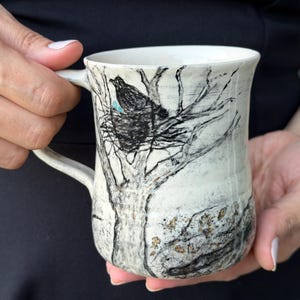 May include: A white ceramic mug featuring a hand-drawn black ink illustration of a bird sitting in a nest within a bare tree. The design includes a small blue accent on the bird and subtle gold-colored floral details near the base of the mug.