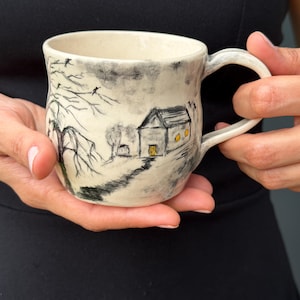 Puede incluir: Una taza de cerámica con base color crema que presenta una ilustración a tinta negra de una casa, un árbol desnudo y un camino. La casa tiene ventanas y una puerta amarillas. La taza tiene un asa curva y un borde ligeramente irregular.
