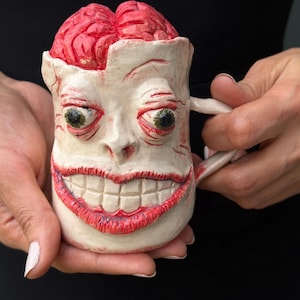 May include: A ceramic mug shaped like a human head with a wide, toothy grin and bulging green eyes. The top of the mug features a textured, bright red exposed brain. The face is off-white with red detailing around the eyes and mouth.