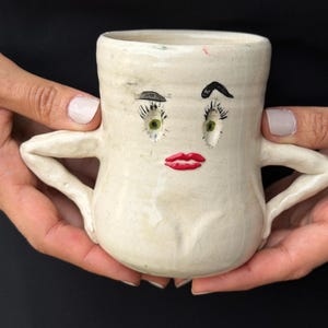 May include: A cream-colored ceramic mug featuring a hand-sculpted face with green eyes, long black eyelashes, arched eyebrows, and bright red lips. The mug has two side handles shaped like arms with hands resting on the hips of the vessel.