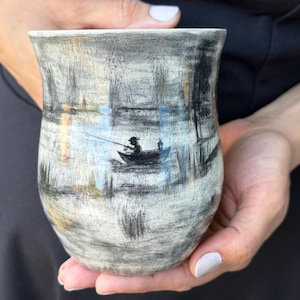 Könnte beinhalten: Ein Keramikbecher mit strukturierter, cremeweißer und grauer Oberfläche, der eine handgezeichnete Illustration einer Person in einem kleinen Boot beim Angeln auf einem See zeigt. Das Kunstwerk enthält dunkelgraue Striche, die Wasser, Schilf und die Silhouette einer Figur mit Angelrute darstellen und ein skizzenhaftes Aussehen auf der gewölbten Oberfläche erzeugen.