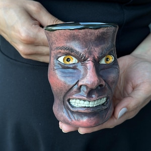 May include: A ceramic mug sculpted in the shape of a human face with a dark reddish-brown finish. The face features intense yellow eyes, furrowed brows, and a bared-teeth expression. The mug has a smooth, dark rim and a textured, realistic skin-like surface.
