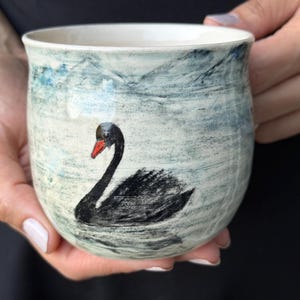 Może przedstawiać: Ceramiczny kubek o zaokrąglonym kształcie z ręcznie malowanym czarnym łabędziem o jaskrawopomarańczowym dziobie. Tło kubka ma teksturowane, jasnoniebiesko-białe wykończenie, które imituje wygląd wody z subtelnymi zmarszczkami na powierzchni.