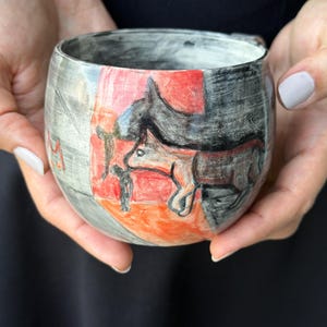 Könnte beinhalten: Ein abgerundeter Keramikbecher mit einem handgemalten Design, das ein schwarz-weiß skizziertes Fuchsmotiv vor einem leuchtend roten und orangefarbenen rechteckigen Hintergrund zeigt. Der Becher hat eine texturierte, gräuliche Oberfläche.