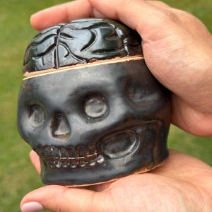 May include: A black ceramic lidded jar shaped like a human skull with a brain-textured lid. The surface has a glossy finish with visible terracotta-colored clay edges where the lid meets the base, featuring carved eye sockets and teeth.
