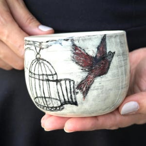 Może przedstawiać: Biały ceramiczny kubek z ręcznie rysowaną czarną ilustracją przedstawiającą otwartą klatkę dla ptaków wiszącą na gałęzi oraz ciemnoczerwonego ptaka wylatującego z klatki. Kubek ma teksturowaną, złamany biały kolor powierzchni z widocznymi pociągnięciami.