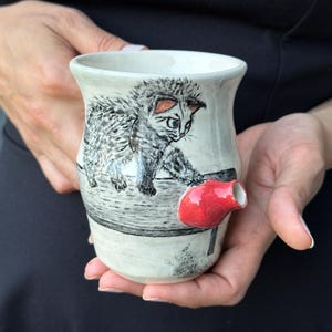 May include: A ceramic mug with a light gray glaze featuring a detailed black ink illustration of a fluffy kitten walking along a ledge. The mug includes a small, protruding, bright red sculptural element shaped like a bird or small animal.
