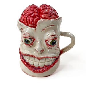 May include: A ceramic mug shaped like a human head with a wide, toothy grin and bulging green eyes. The top of the mug features a sculpted, glossy red brain exposed at the crown. The surface is off-white with red detailing around the mouth and eyes.