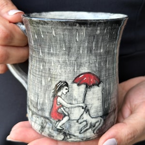 May include: A ceramic mug with a textured gray finish featuring a hand-painted scene of a person in a red dress holding a red umbrella over a small dog during a rainstorm. The mug has a cylindrical shape with a handle and a dark rim.