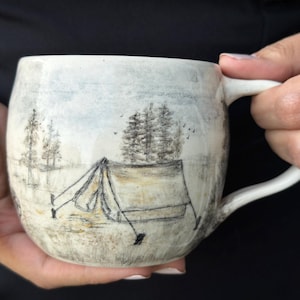 May include: A ceramic mug with a light, speckled off-white glaze featuring a hand-drawn illustration of a camping tent nestled among pine trees. The artwork is rendered in muted black and brown ink tones, depicting a serene outdoor landscape.