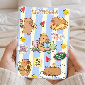 May include: A clear phone case featuring a repeating pattern of blue and yellow vertical stripes. The case is decorated with cartoon capybaras and food items like pizza, watermelon, and ice cream. Text includes "CAPYBARA", "Bookish Baby", and "Emotional Support Kindle".