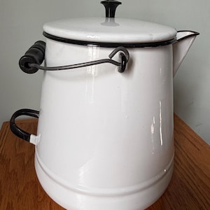 May include: White enamelware coffee pot with a black handle and lid knob. The pot has a curved spout and a wire handle. The rim and handle are trimmed in black. A classic kitchenware item.