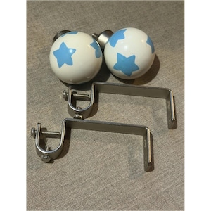 May include: Two spherical white curtain finials featuring a light blue star pattern, paired with two silver-toned metal L-shaped curtain rod brackets. Each bracket includes a circular mounting ring with adjustable screws for securing the rod in place.