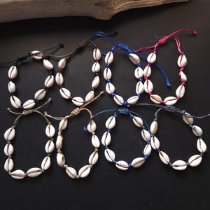 Op de afbeelding: Acht verstelbare armbanden met witte kaurischelpen aan gekleurde koorden in zwart, bruin, blauw, roze, beige en grijs. Elke armband heeft een schuifknoopsluiting en is gerangschikt in twee rijen van vier op een donkere ondergrond.