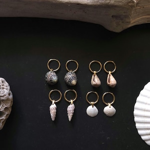Peut inclure: Huit paires de boucles d'oreilles en coquillages avec des cerceaux dorés sont présentées sur une surface noire. Les boucles d'oreilles présentent différents types de coquillages, y compris des coquillages tachetés, coniques et blancs plats. Un morceau de bois flotté et une grande coquille sont également visibles.