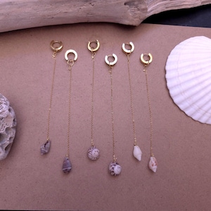 以下が含まれることがあります： 小さな貝殻のペンダントが付いた、6つのゴールドカラーのチェーンピアス。 ピアスは茶色の表面に展示されており、背景には白い貝殻と流木があります。 ピアスは繊細でミニマリストなデザインです。