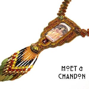 May include: A gold beaded necklace with a rectangular pendant featuring a vintage image of a couple. The pendant is surrounded by gold beads and has a red gemstone accent. The necklace has a long tassel with gold and black beads and gold leaf accents. The text "Moët & Chandon" is printed below the necklace.