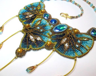 Può includere: Una collana di perline con un design a farfalla in tonalità di blu, oro e argento. La collana ha una lunga catena pendente con più perline.