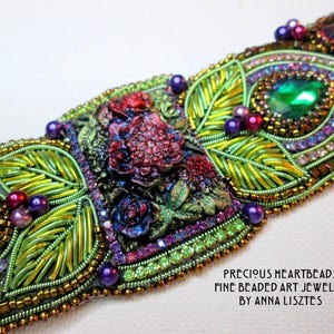 May include: A handcrafted beaded art jewelry piece with intricate floral and leaf designs. The piece features green, purple, and red beads, along with a large green gemstone. The text "PRECIOUS HEARTBEADS FINE BEADED ART JEWELRY BY ANNA LISZTES" is visible.